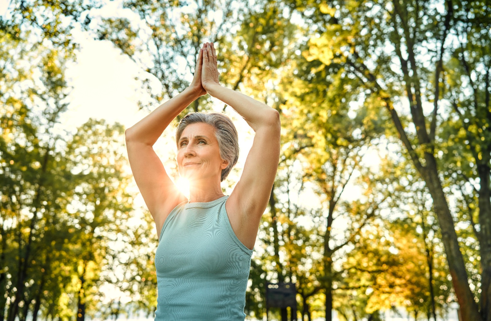 Older woman doing yoga in morning sunlight, surrounded by trees.