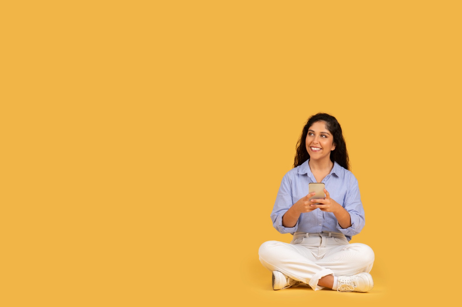 Young Indian woman smiling while holding a cup, sitting cross-legged against a bright yellow background, symbolizing confidence and hormonal balance.