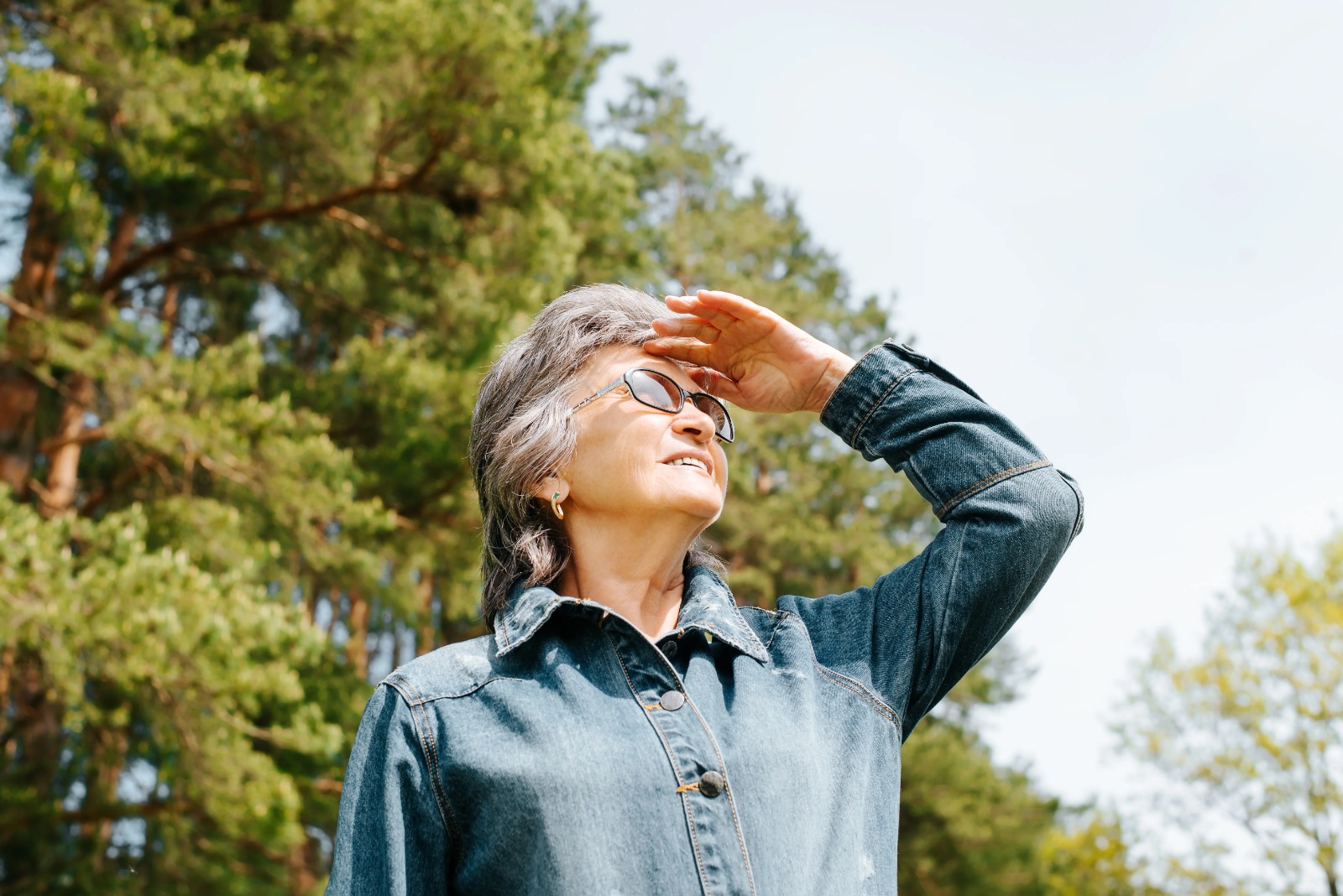 Older woman shielding her eyes from bright sunlight while wearing sunglasses outdoors.