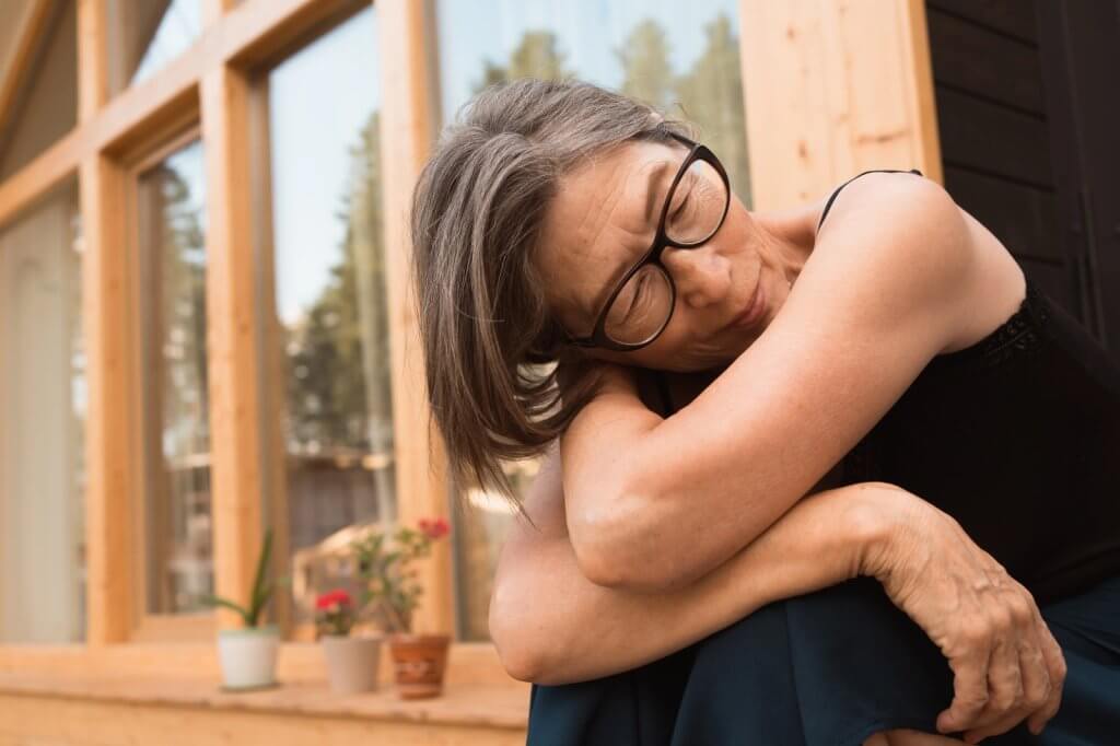 Senior Asian woman with grey hair and glasses resting peacefully outdoors, eyes closed and arms wrapped around knees in this blog on menopause mood swings versus depression.