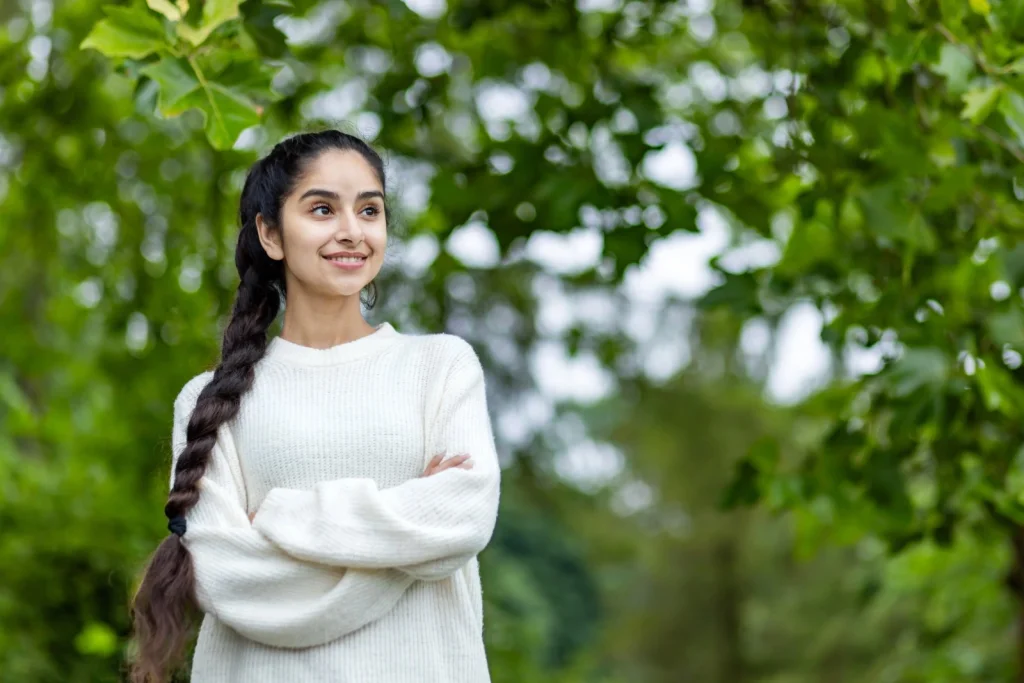 A happy young Indian woman with long braided hair wearing a cozy white sweater, smiling confidently outdoors with lush green trees in the background, symbolizing health and positivity