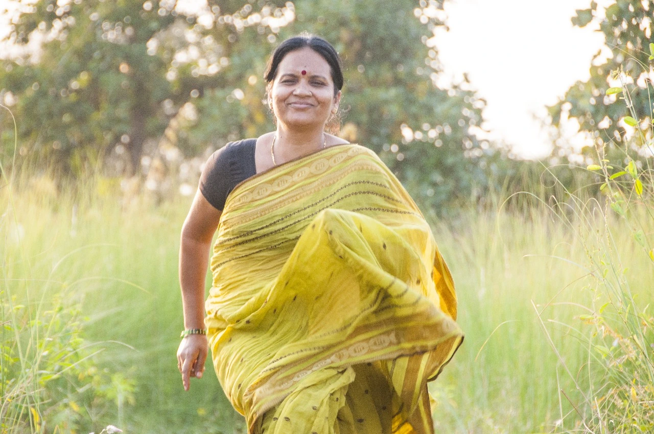 Smiling middle-aged woman in a yellow saree walking outdoors in a green field during sunset, radiating energy and wellness.